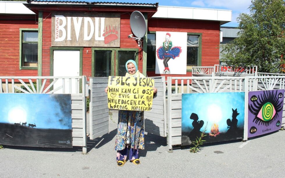 Julie po­se­rer ut­en­for den ned­lag­te pu­ben Biv­du i Kar­as­jok, vegg i vegg med Sá­gat-kon­to­ret. På pla­kat­en hen­nes står det blant an­net at «Hvi­le­da­gen er lør­dag. Hal­le­lu­ja!». Julie er ty­de­lig på at det var en feil av kir­ka å flyt­te hvi­le­da­gen til søn­dag.
 Foto: Elise Embla Scheele