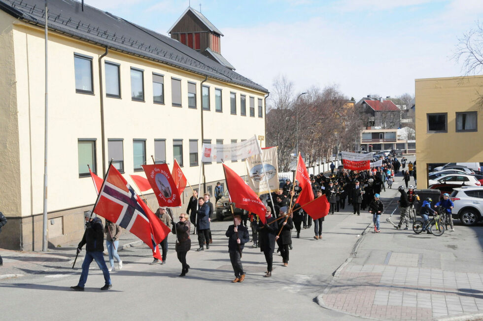 Her er toget på vei til Råd­hus­plas­sen med horn­mu­sik­ken og tromme­orkes­ter samt flagg­borg i front. Del­ta­kerne var i år færre enn tid­lig­ere år, og det er en stor andel eldre og gamle som fører tra­di­sjo­nen vid­ere.
 Foto: Hallgeir Henriksen