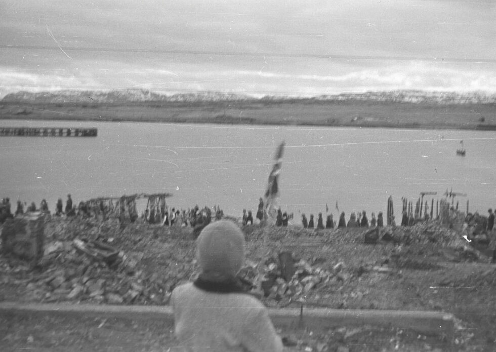 Mennesker i alle aldre står ved fjorden i Vadsø, og norske flagg bæres med stolthet. I forgrunnen står et barn med ryggen til, og ser mot feiringen. Naturen er majestetisk, men landskapet rundt bærer fortsatt preg av krigens herjinger.
 Foto: Leif Hauge. Finnmark Fylkesbibliotek