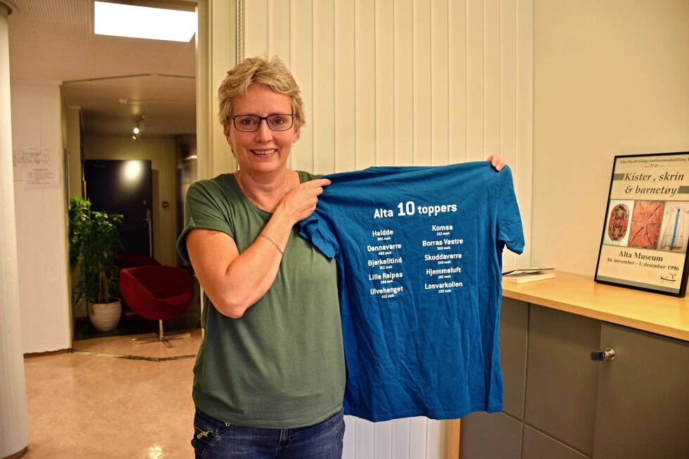 Rådgiver på kulturavdelingen i Alta kommune, Aase-Kristin H. Abrahamsen viser frem t-skjorten for ti-toppers turer, som nå også får navnene på toppene på samisk.
 Foto:  Ávvir, Marit Elin Kemi